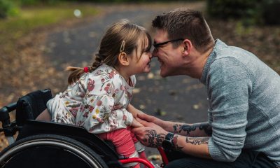 A man smiling and facing a young girl in a wheelchair, also smiling, on a nature path